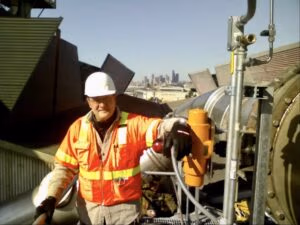 Rod Gravley, dressed in a tan shirt, an orange safety vest, black work gloves, and a white hard hat, leans casually against a piece of industrial equipment during a site visit.